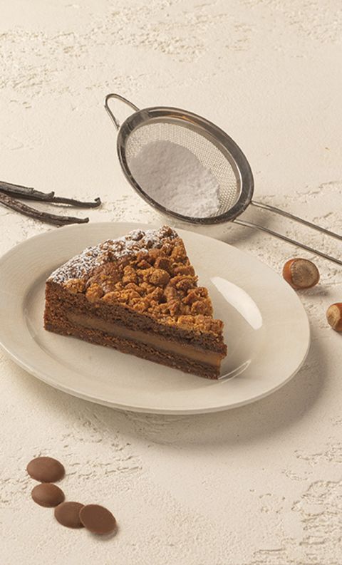 Fetta di torta al cioccolato con crumble e ripieno al caramello su piatto bianco, con nocciole e stecche di vaniglia