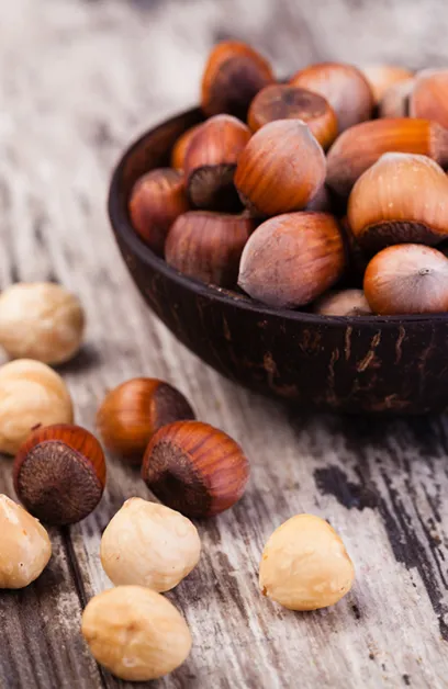 Nocciole intere con guscio in una ciotola di legno scuro, con gherigli sparsi su un tavolo rustico