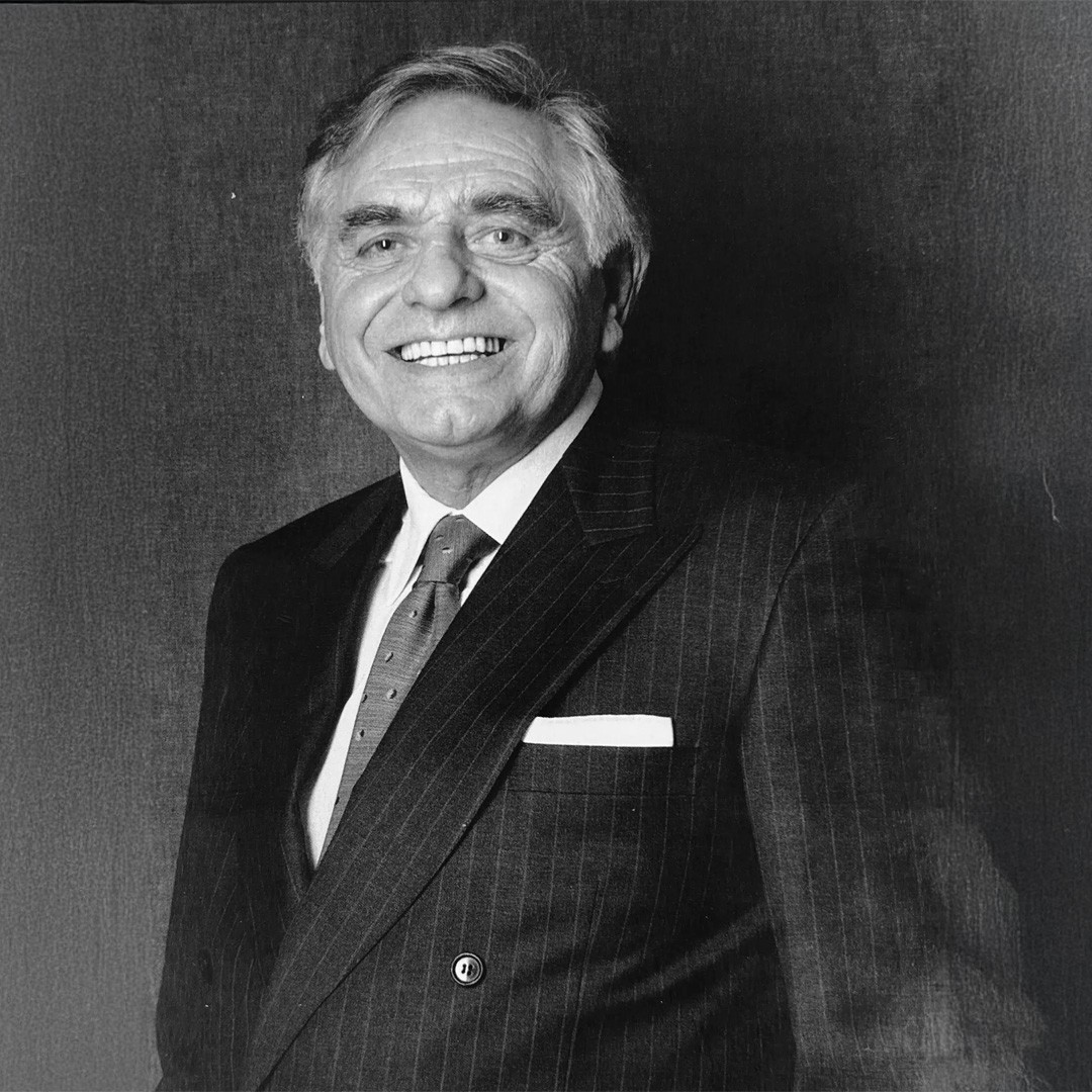 Ritratto in bianco e nero di un uomo anziano sorridente in abito gessato, cravatta a pois e pochette, sfondo scuro
