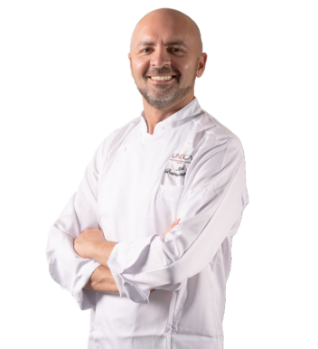 Uomo calvo sorridente con barba, braccia incrociate, giacca da chef bianca con logo ricamato, sfondo neutro