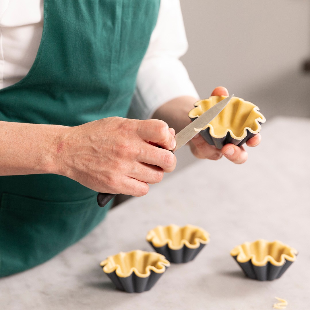 Persona con grembiule verde rifila la pasta in eccesso da uno stampo nero per tartellette con un coltellino.