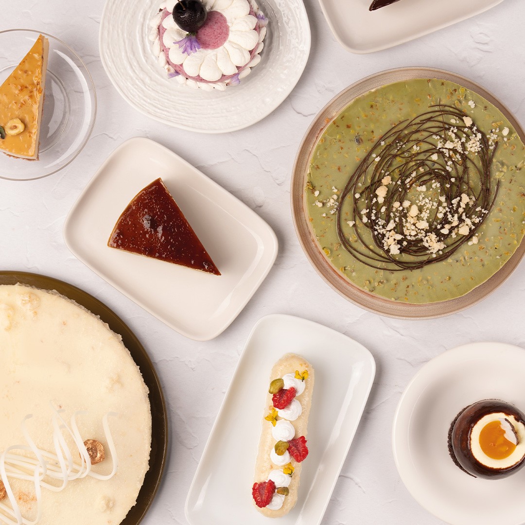 Vista dall'alto di dessert assortiti: cheesecake, flan, crostata al pistacchio, eclair e coppette di mousse al ciocco...