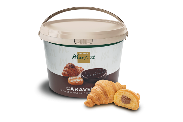 Secchiello beige di crema spalmabile al cioccolato Caravel Master Martini accanto a due croissant, uno farcito di cio...