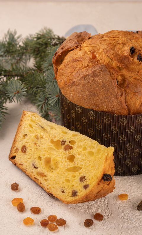Panettone con fetta tagliata, uvetta e canditi, accanto a rami di abete su sfondo chiaro.