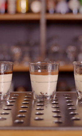 Tre bicchierini con liquore crema a strati e cacao su bancone metallico, bar sfocato sullo sfondo.