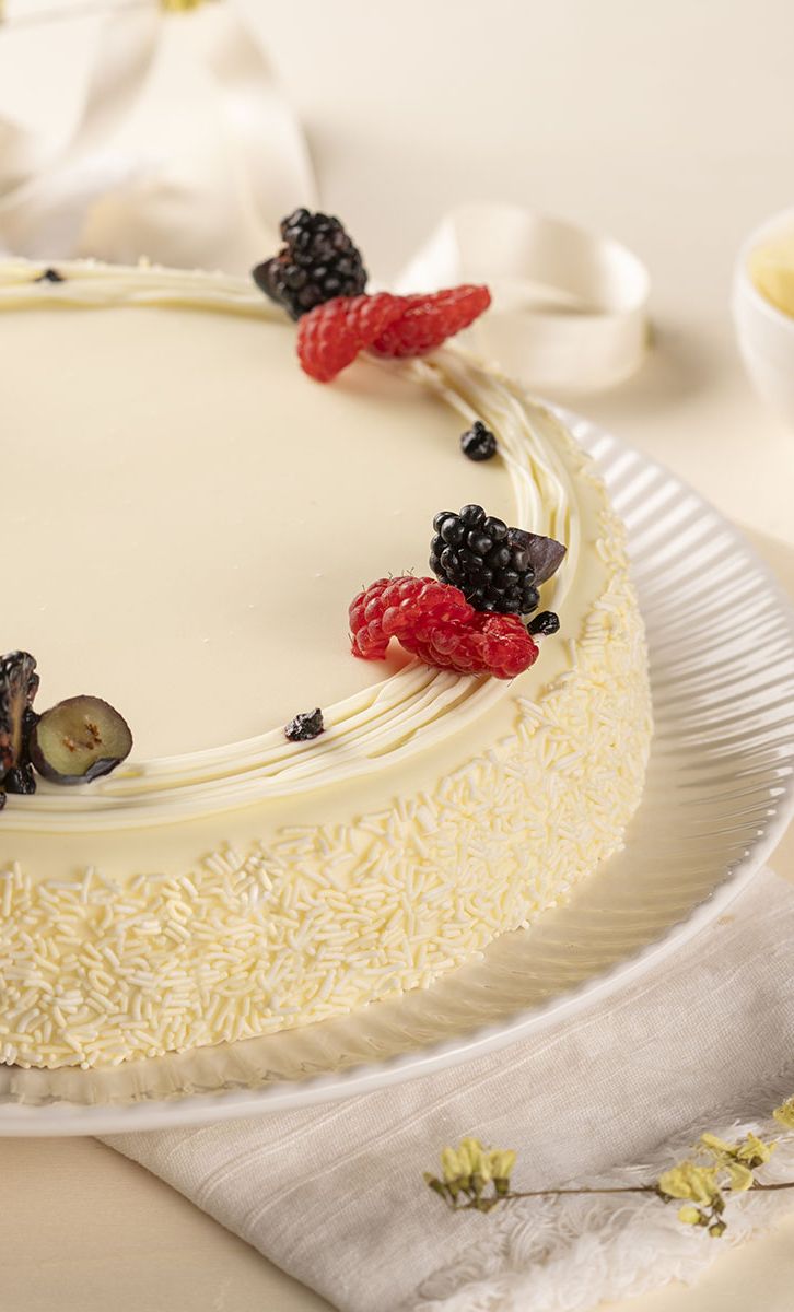 Torta ricoperta di cioccolato bianco con lamponi e more, su piatto con scaglie di cioccolato.