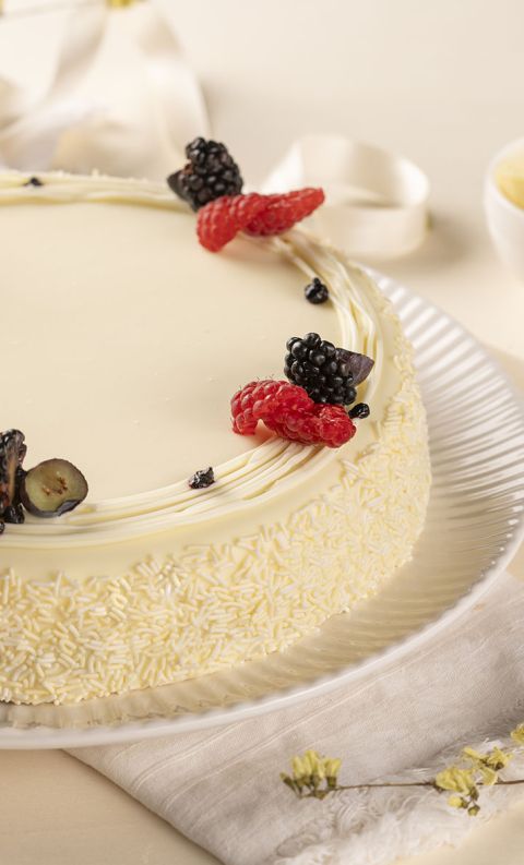 Torta ricoperta di cioccolato bianco con lamponi e more, su piatto con scaglie di cioccolato.