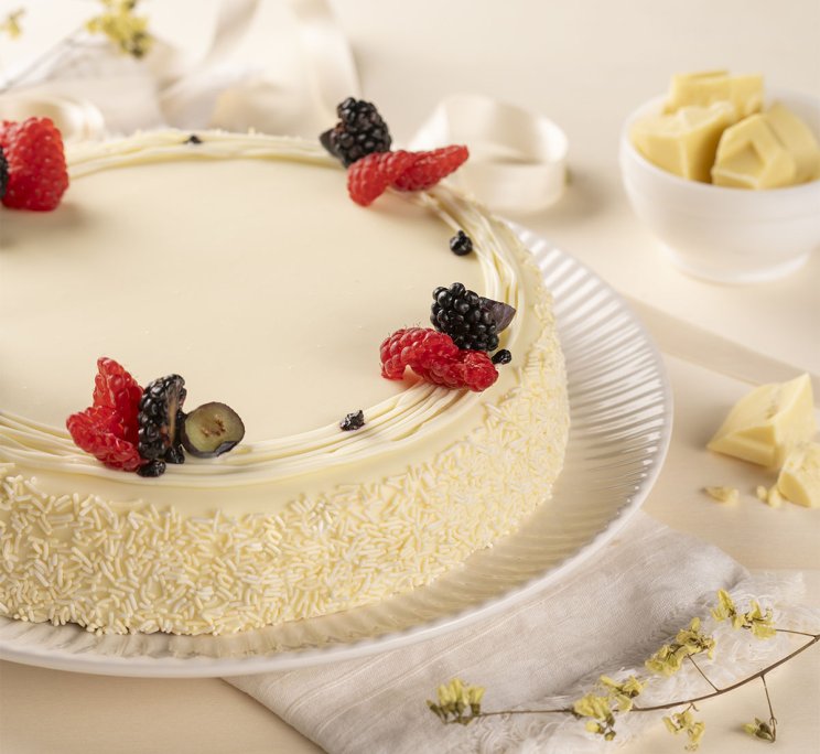 Torta ricoperta di cioccolato bianco con lamponi e more, su piatto con scaglie di cioccolato.