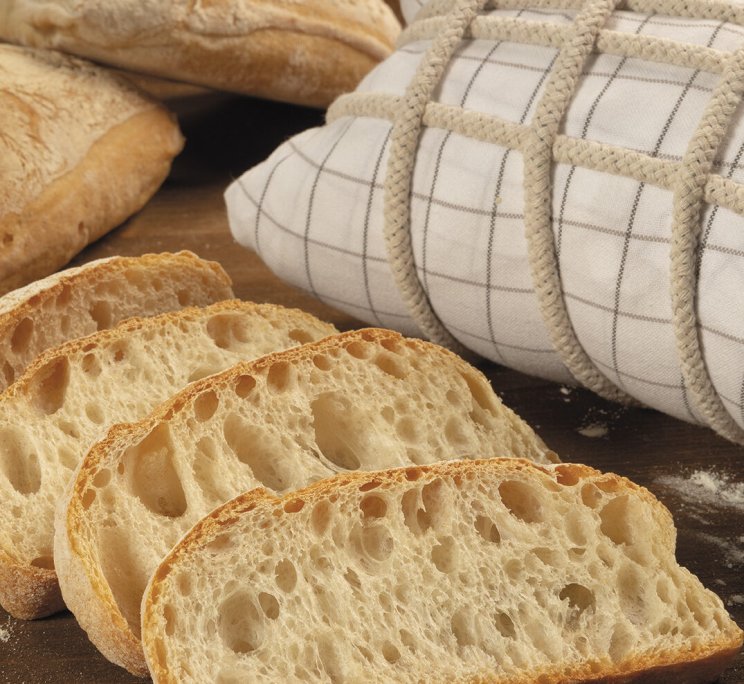 Pagnotte artigianali affettate con mollica soffice, accanto a un pane avvolto in tela a quadretti su asse infarinata