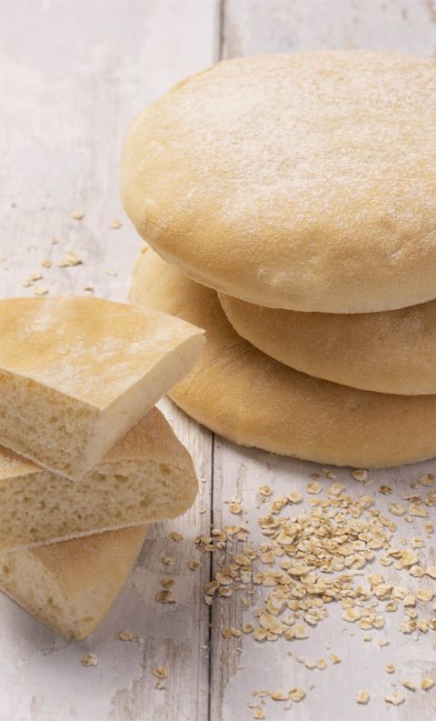 Pane piatto rotondo impilato accanto a fette con interno morbido, avena sparsa su superficie in legno bianco.