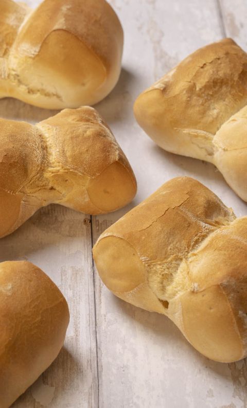 Cinque panini a forma di farfalla dorati, spolverati di farina, su un tavolo rustico in legno bianco.