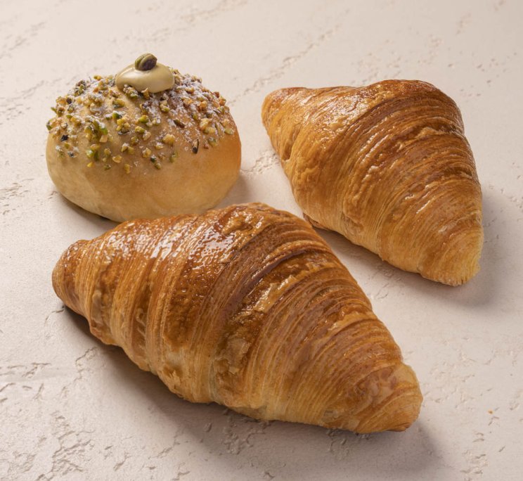Due croissant dorati accanto a una pasticceria rotonda con granella di pistacchio, crema e zucchero a velo.