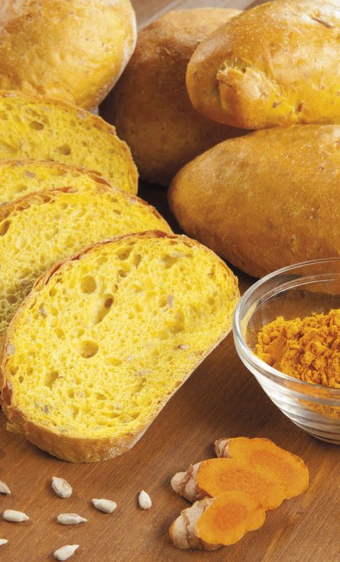 Pane alla curcuma a fette con mollica gialla, pagnotte intere, ciotola di curcuma in polvere, semi di girasole ed erb...