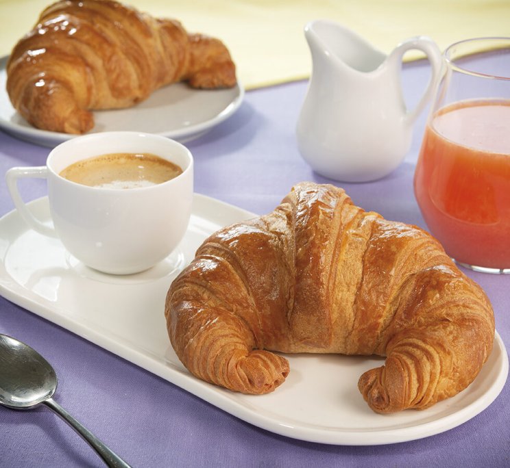 Colazione con croissant dorato su un piatto, tazza di caffè, secondo croissant, panna e succo di frutta.