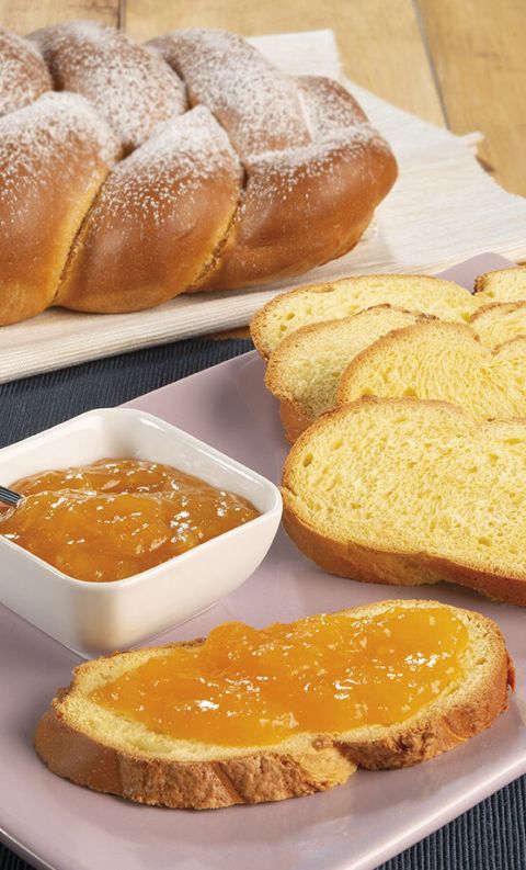 Treccia di pan brioche a fette su piatto rosa, una con marmellata di albicocche accanto a una ciotolina di confettura