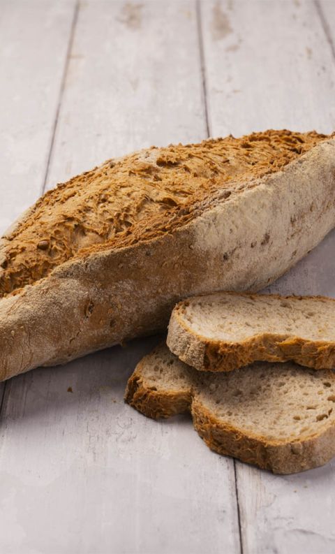 Pagnotta artigianale oblunga con crosta farinata e screpolata, accanto a tre fette su legno bianco.