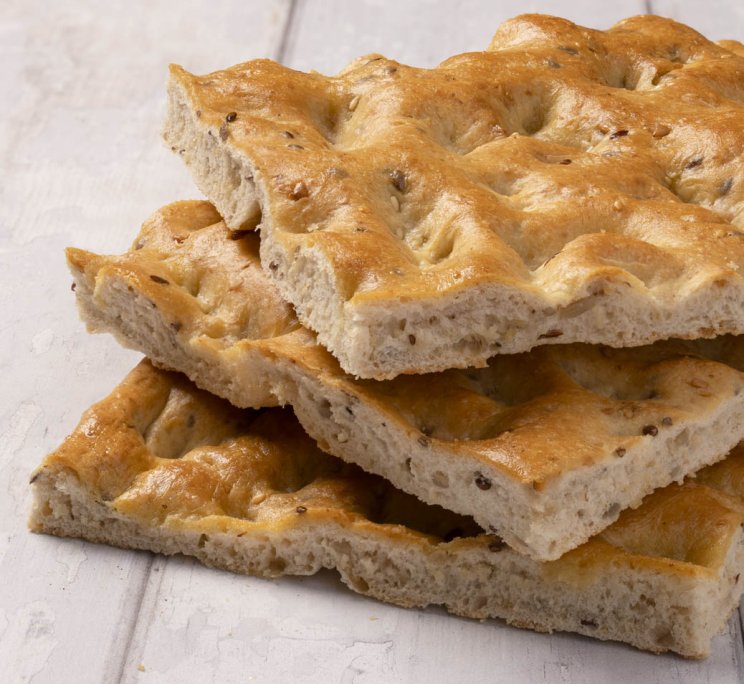 Fette impilate di focaccia dorata con crosta a fossette e semi, su superficie di legno bianco.