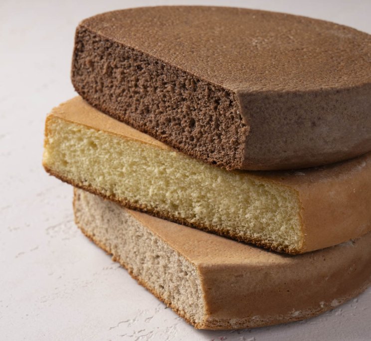 Tre strati rotondi di pan di Spagna al cioccolato, pistacchio e vaniglia su superficie bianca.