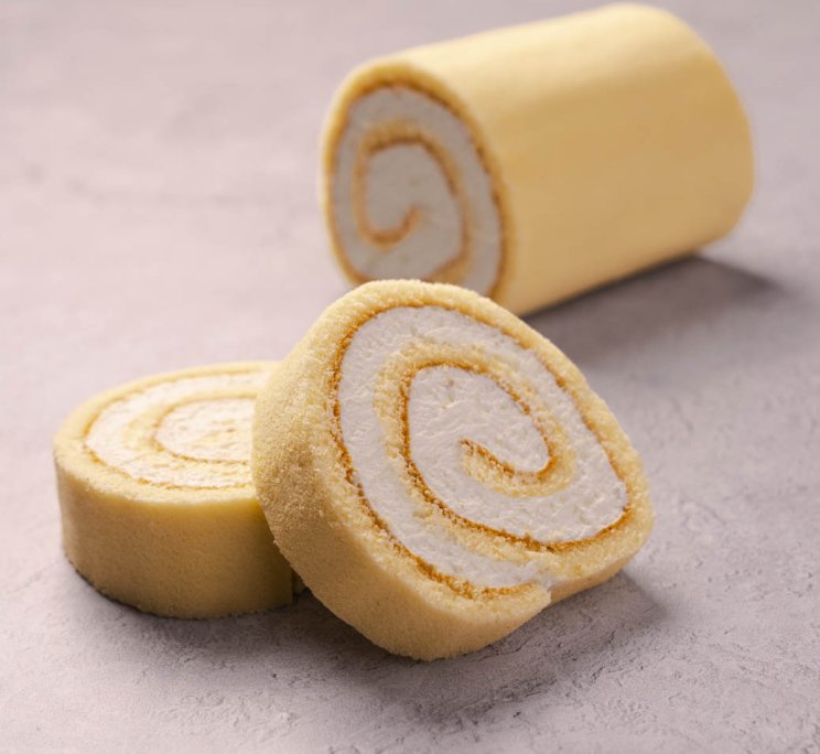 Rotolo di pan di spagna giallo affettato con crema bianca a spirale, su superficie grigia testurizzata.