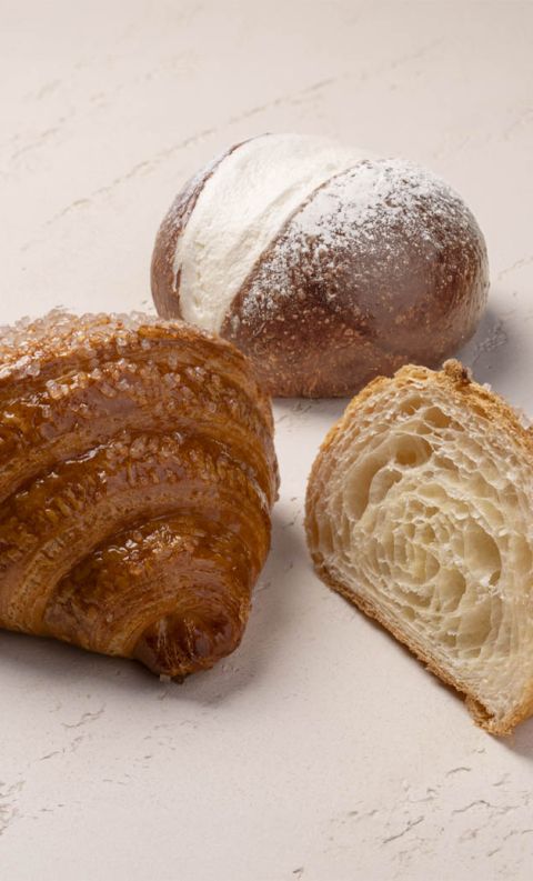 Croissant con zucchero tagliato a metà accanto a un rollè spolverato di zucchero a velo su superficie chiara.