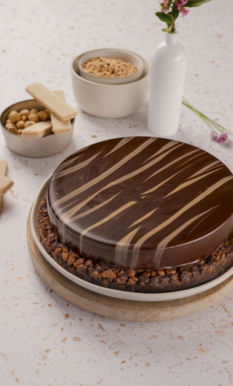 Torta rotonda con glassa a specchio al cioccolato e crosta di frutta secca, su tagliere con wafer, nocciole e fiori