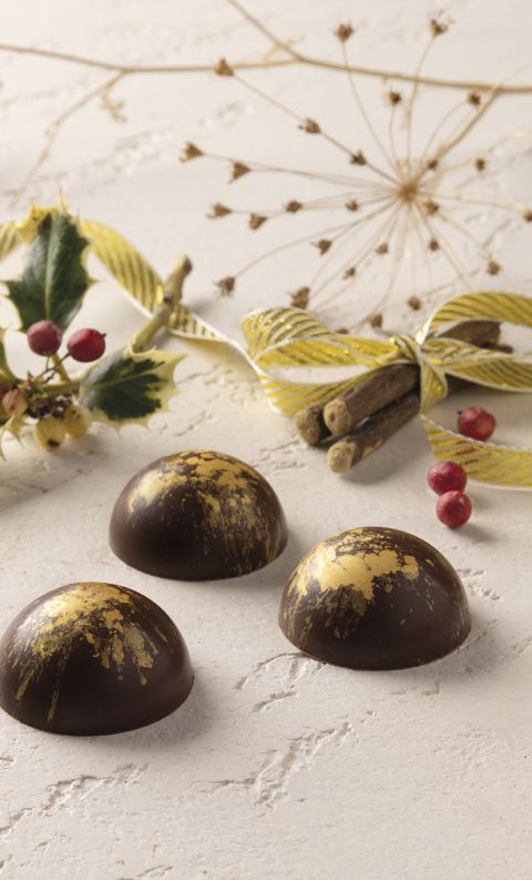 Tre cioccolatini fondenti a cupola con foglia d'oro, circondati da bacche di agrifoglio e stecche di cannella