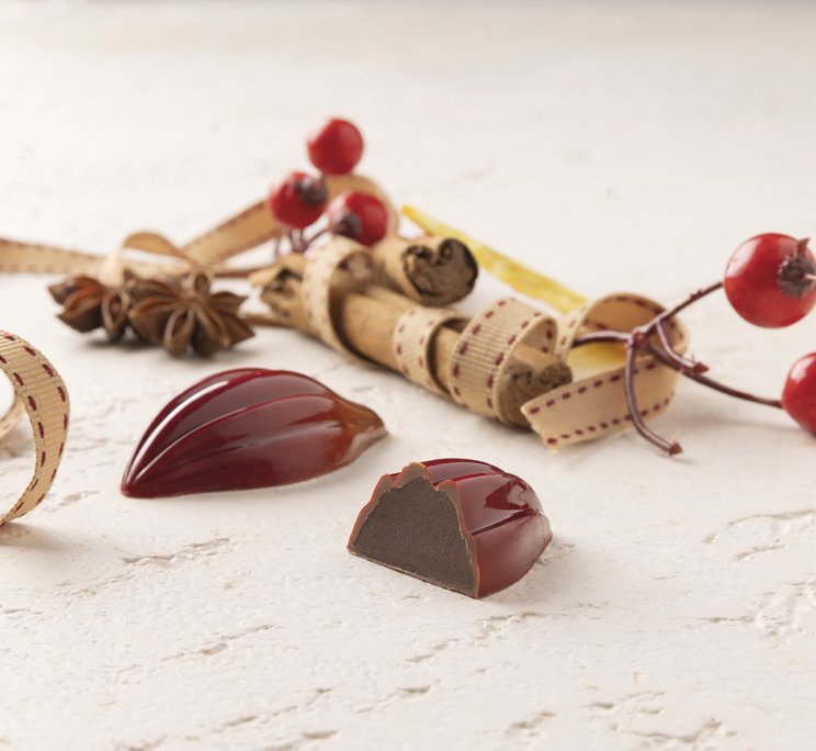 Due cioccolatini rossi lucidi con ripieno scuro, uno tagliato a metà, accanto a cannella, anice stellato e bacche rosse.