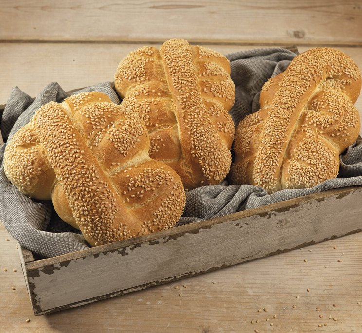 Panini intrecciati con semi di sesamo in un vassoio di legno foderato di tessuto grigio su tavolo rustico.