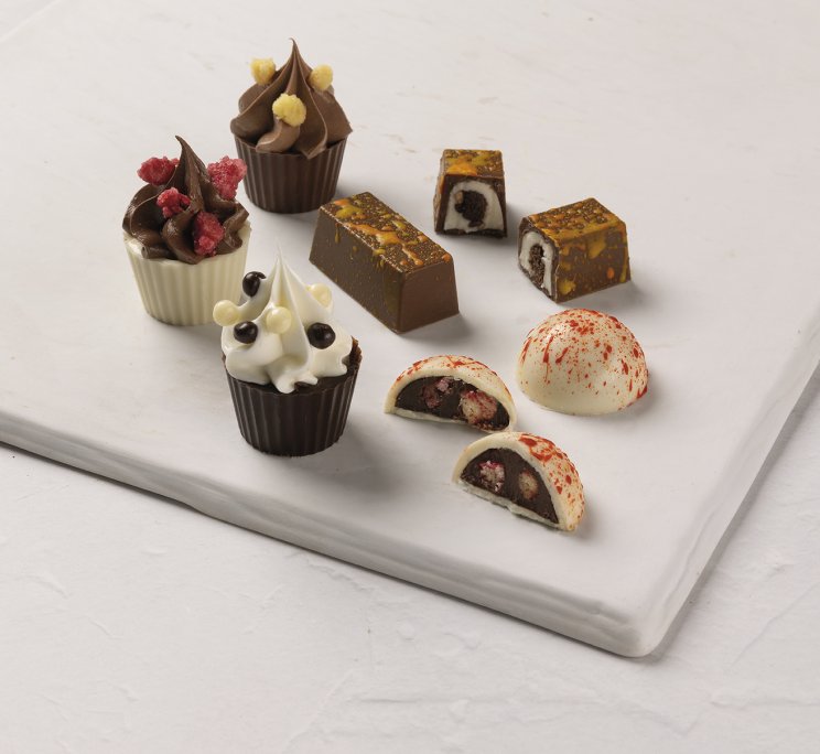 Assortimento di cioccolatini gourmet, con coppette alla crema e praline ripiene, su lastra di marmo bianco.