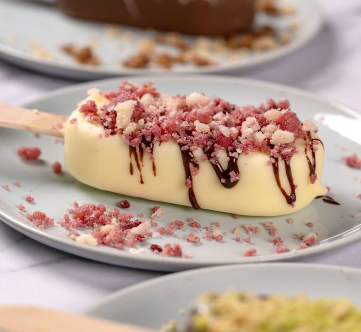 Stecco gelato ricoperto di cioccolato bianco, con colatura di cioccolato fondente e granella di frutta liofilizzata r...