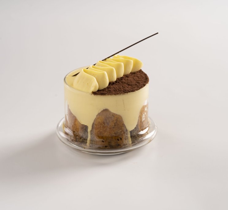 Dessert rotondo stile tiramisù con ciuffi di crema, cacao in polvere e stecco di cioccolato su piatto di vetro.
