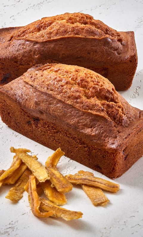 Due filoncini di banana bread dorati con chips di banana disidratata disposte davanti, su superficie bianca.