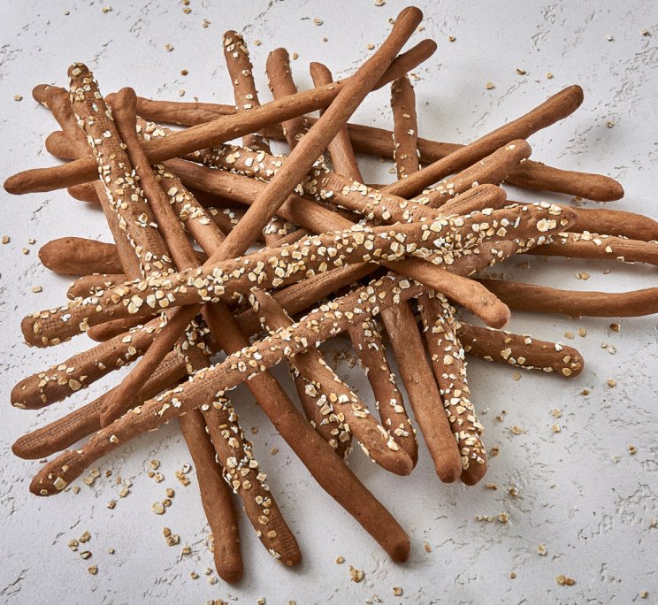 Mucchio disordinato di grissini integrali con avena su una superficie bianca testurizzata.