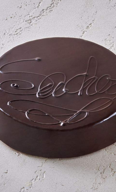 Torta rotonda al cioccolato fondente con glassa lucida e scritte decorative, su superficie bianca testurizzata