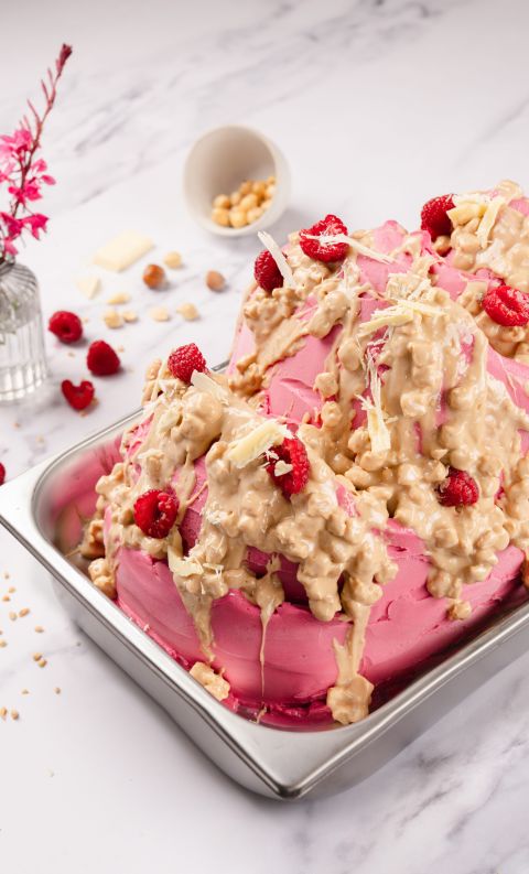 Torta rosa glassata in vassoio metallico, con granella di nocciole caramellate, scaglie di cioccolato bianco e lamponi